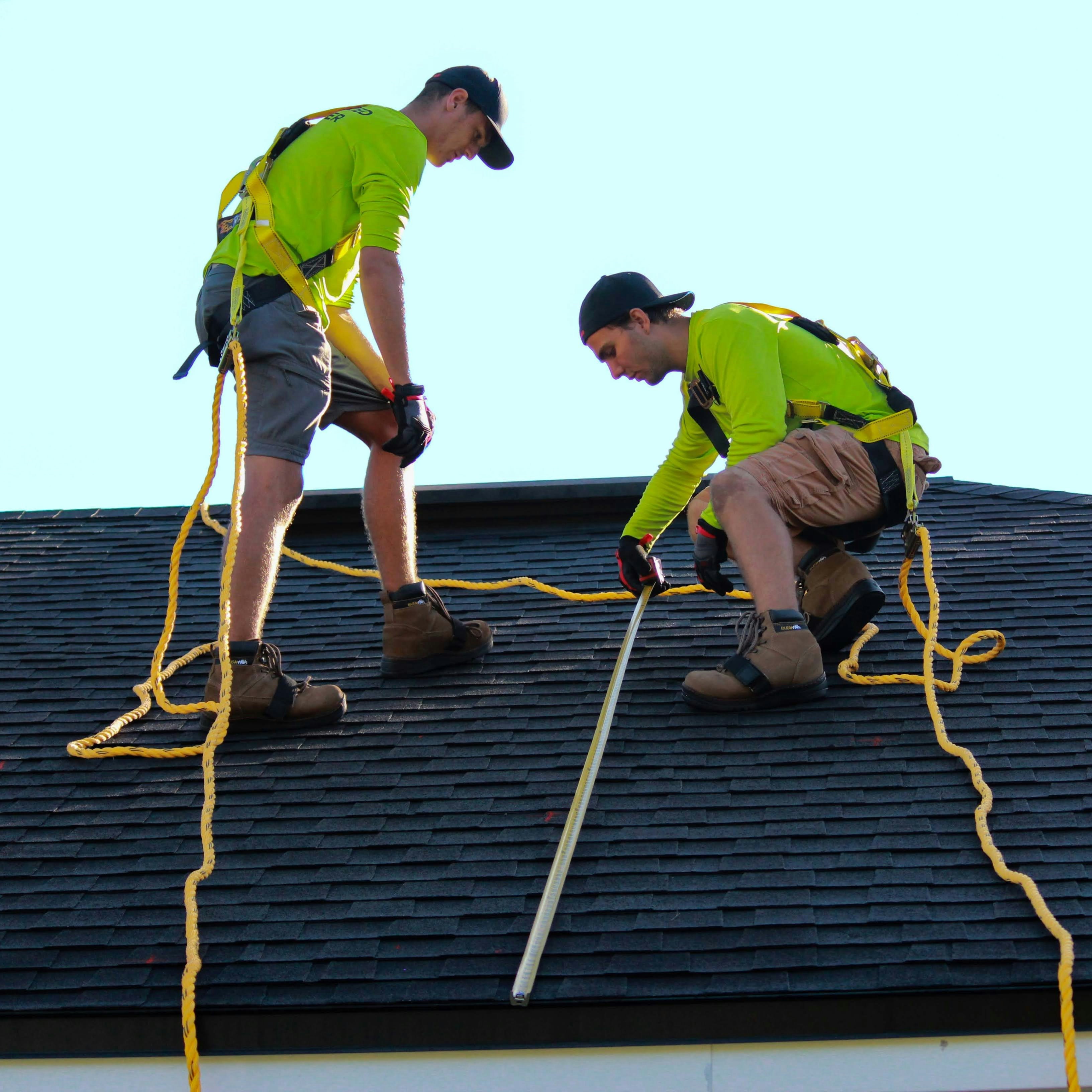 Two roofing professionals in safety harnesses measuring shingles on a residential roof